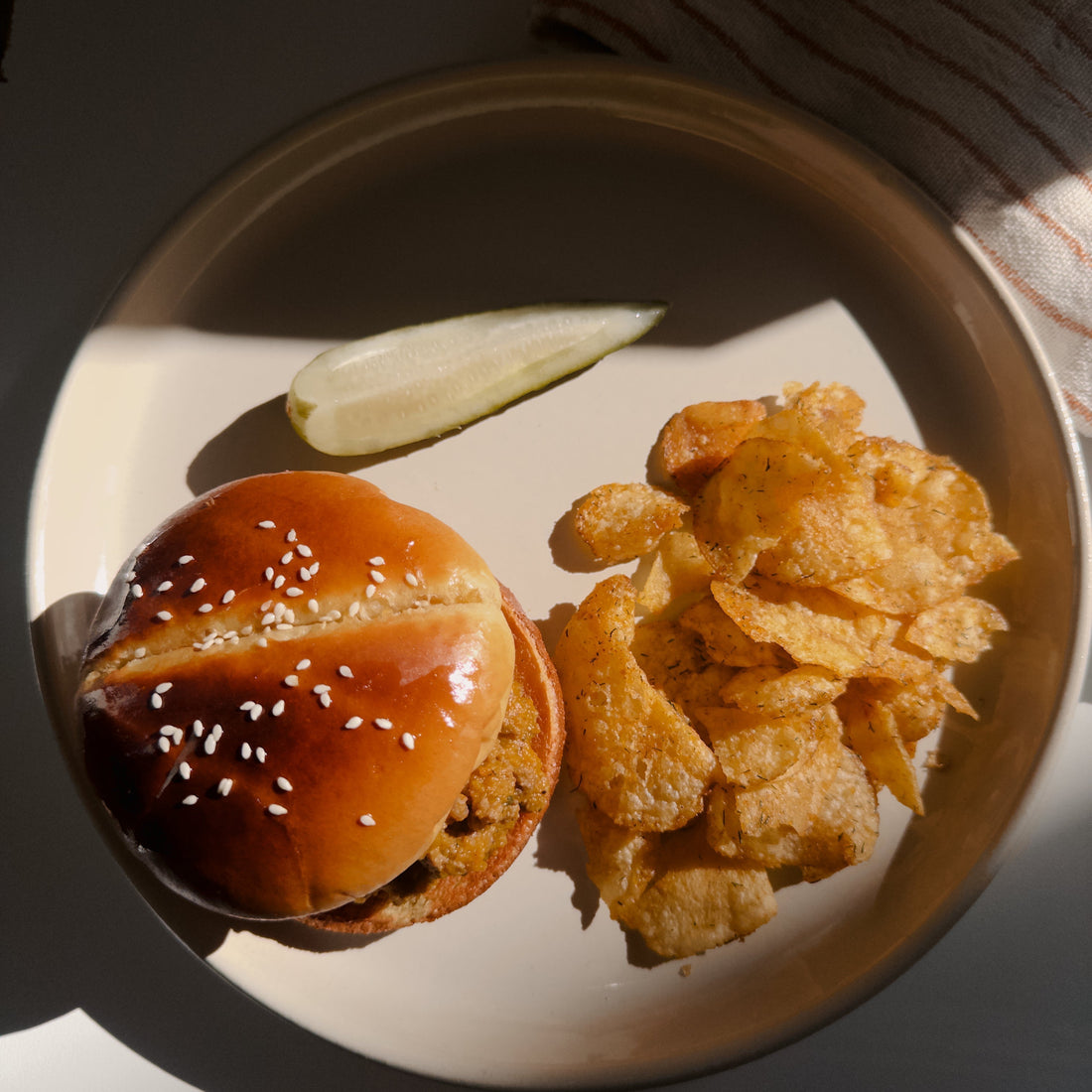 A CARINARA Sloppy Joe on a toasted sesame brioche bun, served with kettle chips and a pickle spear on a round plate.