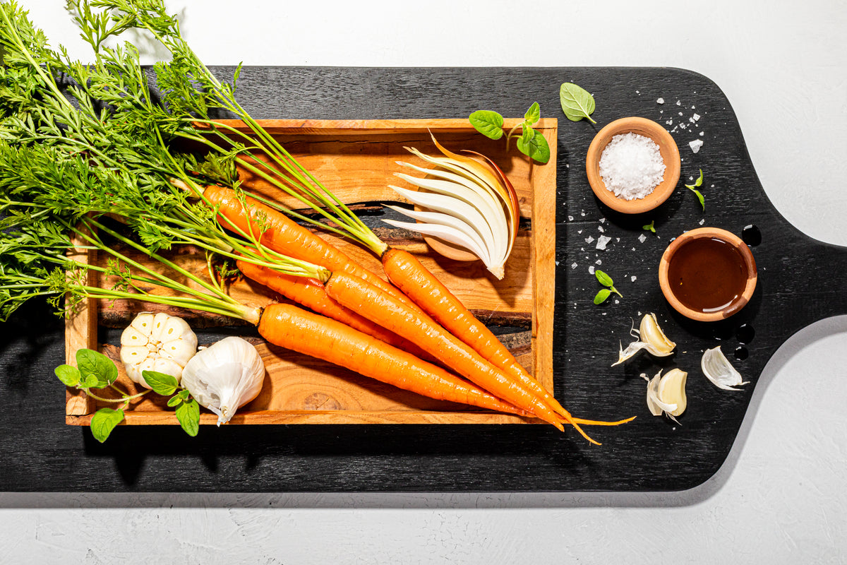 A cutting board displaying the fresh ingredients for CARINARA pasta sauce on a black wood surface. The ingredients displayed include carrots, garlic, onion, salt, oregano, and olive oil