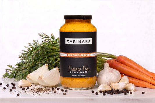 A jar of Carinara Cracked Pepper Tomato Free Pasta Sauce is displayed on a white marble surface surrounded by its raw ingredients, including fresh garlic, whole garlic, carrots, and peppercorns, against a clean white background.