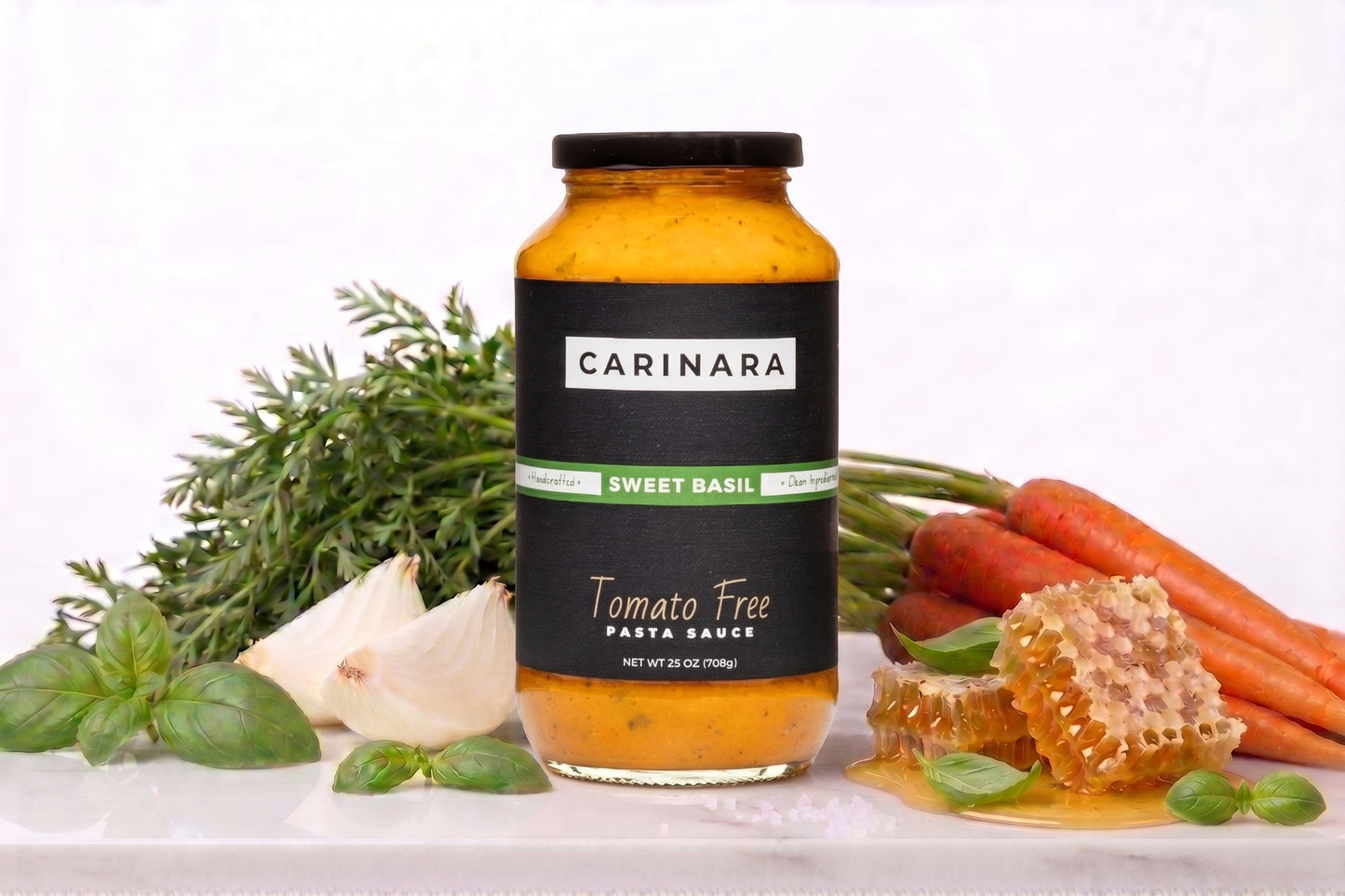 A jar of Carinara Sweet Basil Tomato Free Pasta Sauce is displayed on a white surface surrounded by its natural ingredients, including fresh basil, garlic, carrots, and honeycomb.