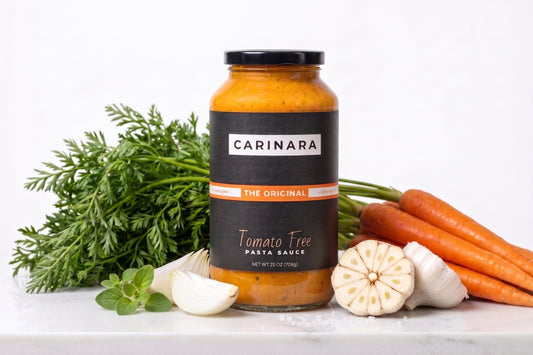 A jar of The Original CARINARA Tomato Free Pasta Sauce stands on a white surface surrounded by fresh ingredients including carrots, garlic, and herbs against a clean white background.